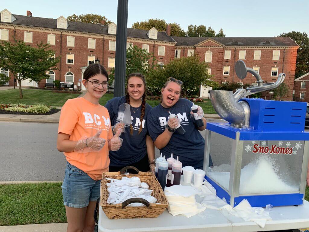 Three girls work a snow cone machine during welcome week on their college campus