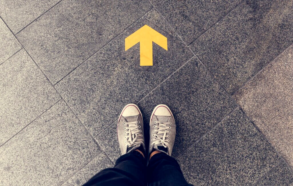 Zoomed in on legs of a person standing with tennis shoes on behind a yellow arrow on the floor