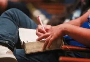 College student with bible in lap taking notes during church service