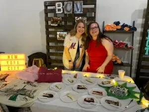 2 Girls pose in front of table with New York City themed items on it including slices of cheesecake