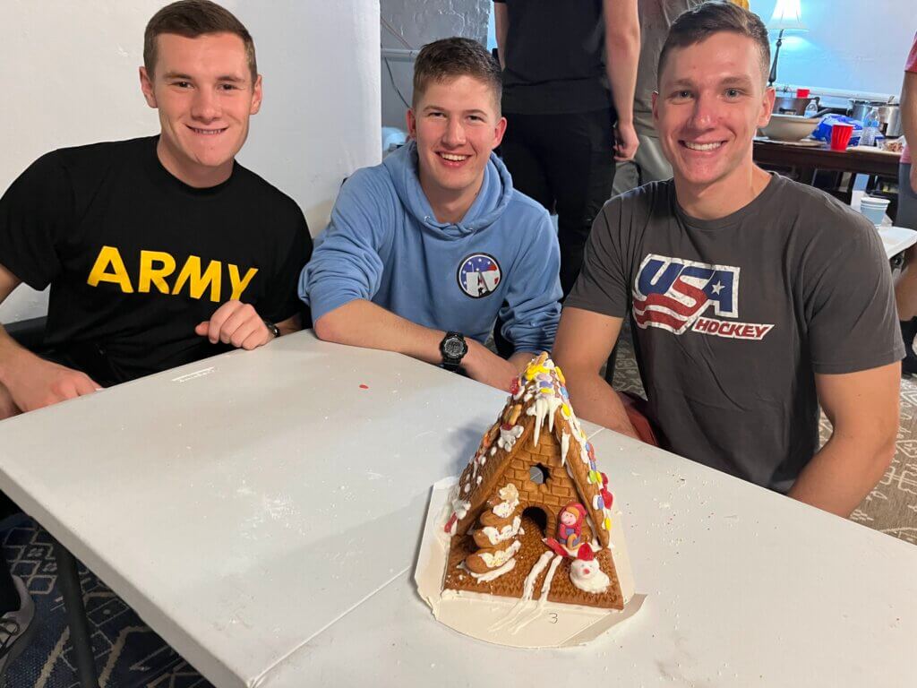 Three college guys pose in front of the gingerbread house they have made.