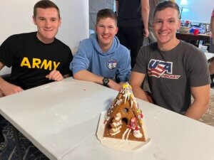 Three college guys pose in front of the gingerbread house they have made.