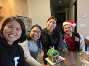 Four co-ed college students pose in front of the gingerbread house they have made.