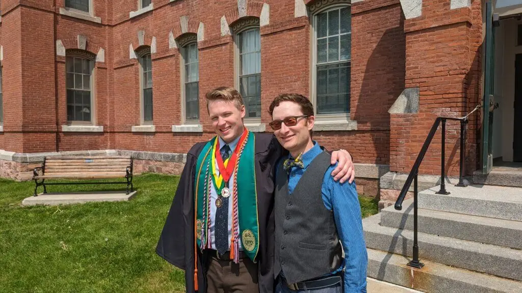 Two students, one in graduation gown with honors cords and medals, pose in front of academic building on college campus