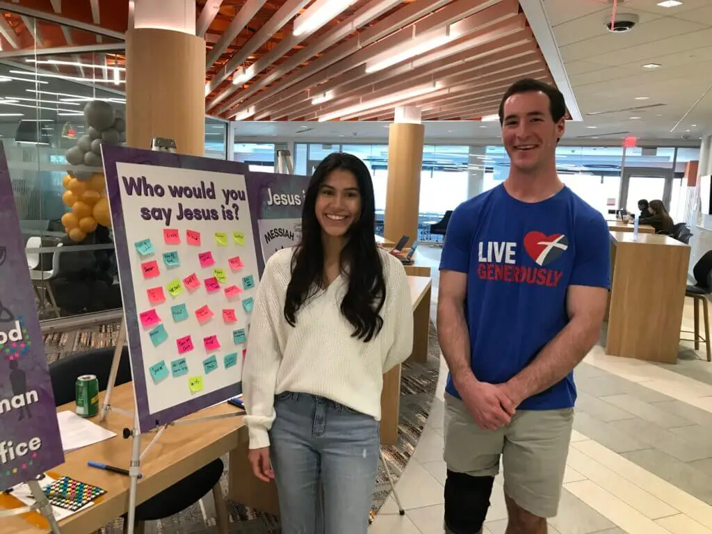 Two college students stand beside sign reading "Who would you say Jesus is?" with sticky note answers on it.