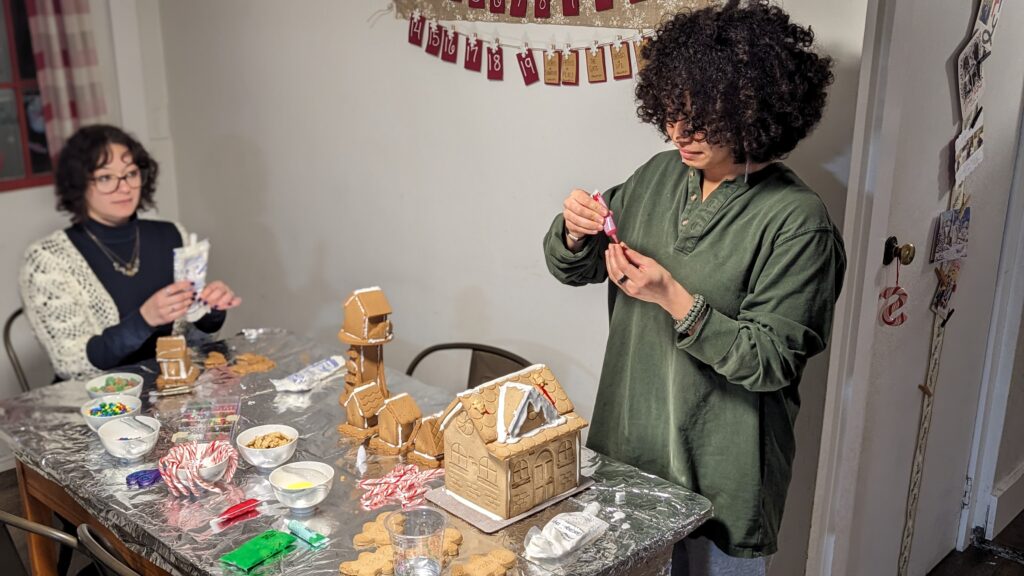 Two college women decorate gingerbread houses at a kitchen table at Christmas time.