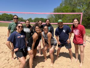 College student athletes pose for a picture on a sand volleyball court with a net in the background.