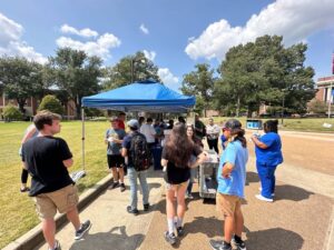 Campus ministry interacting with college students on campus. Standing under and around a blue tent on a campus quad.