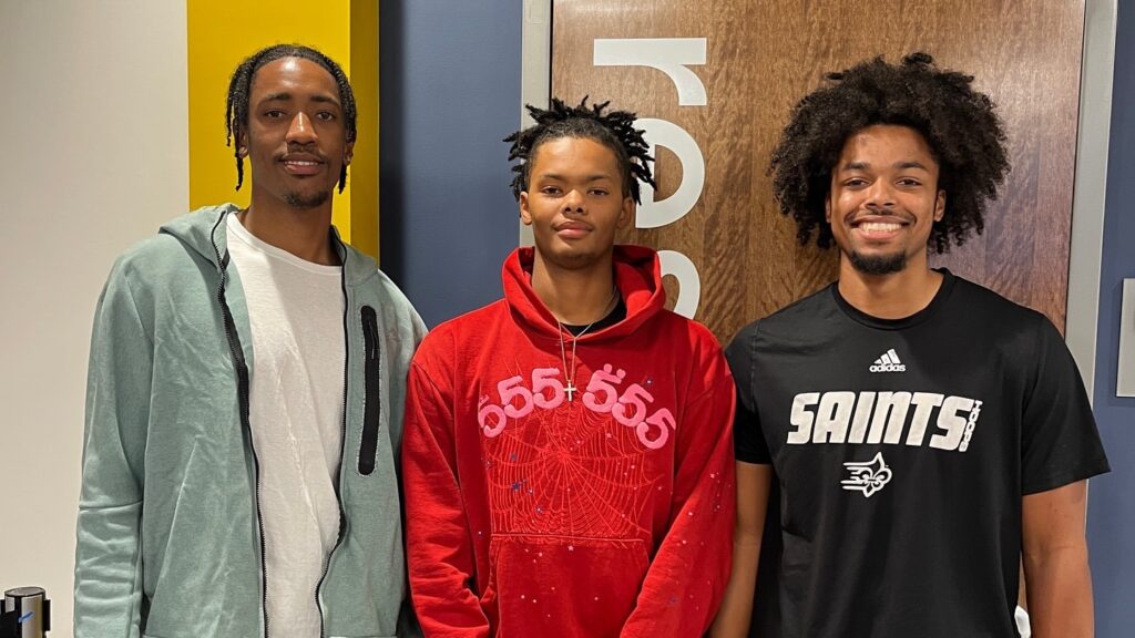 Three college guys pose for a picture wearing a light green zip-up hoodie, a red hoodie with numbers, and a black t-shirt with the word "saints" standing from left to right.