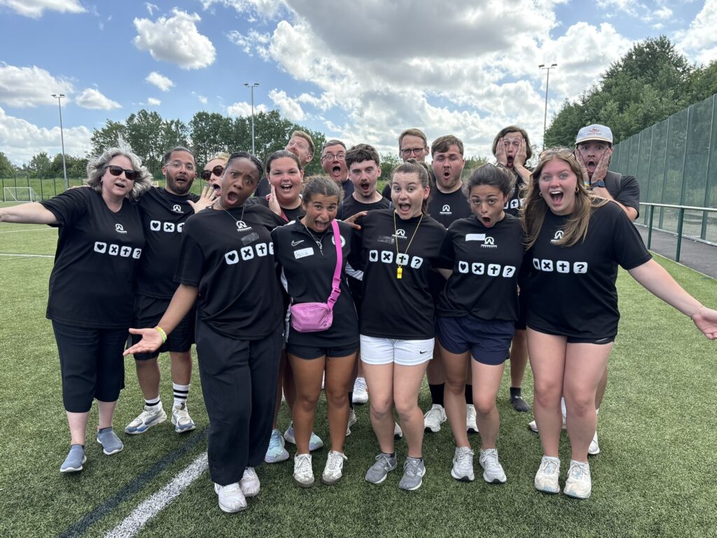 Group of people all in matching black shirts stand outside on a turf field making silly faces.